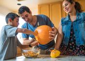 Boy empties pumpkin guts into a bowl while father holds pumpkin. Mother smiles as she looks on at kitchen counter. Photo credit: ltsskin/iStock.