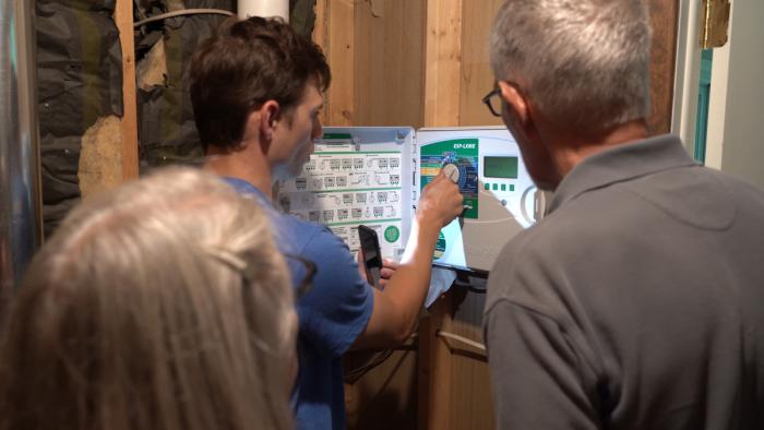 Three people looking at a control panel on a sprinkler system on a garage wall. 