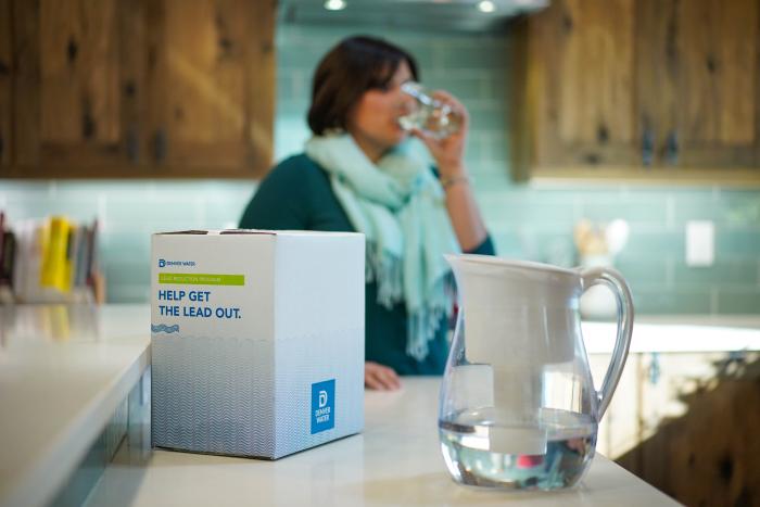 A box with the words "Help get the lead out" sits on a counter, there's a pitcher with a filter that's full of water and a woman, blurred, is in the background drinking a glass of filtered water in her kitchen.