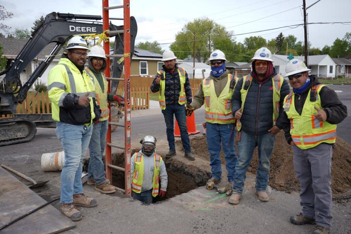 A construction crew of men in hard hats, safety vests and boots, including one man who is in a hole in the street, look up to smile at the camera. Behind them all is a bulldozer and they're in a neighborhood setting with homes. 