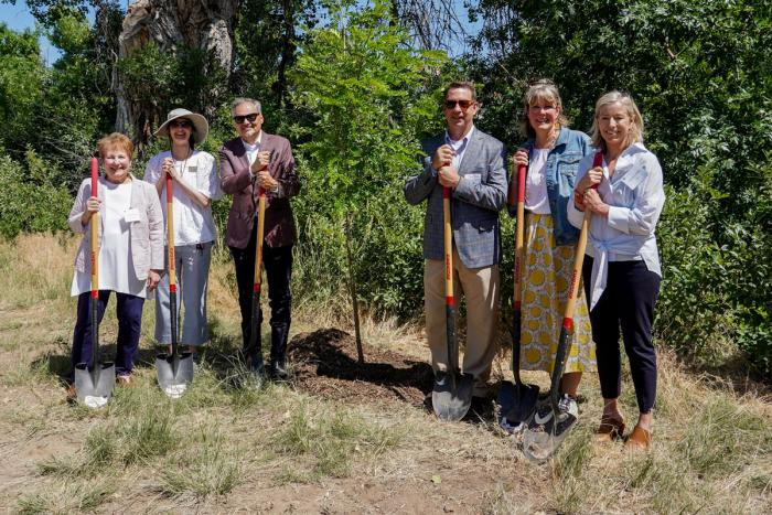 Six people holding shovels stand next to a tree they have just helped to plant. They're smiling in the sunshine and proud of the work they've done.