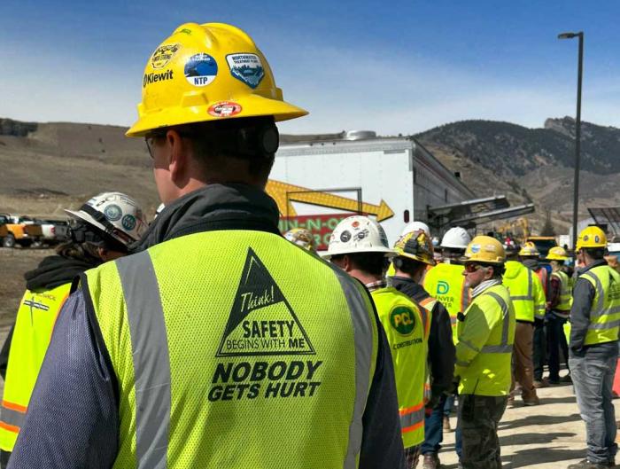 There's a line of people, all with their back to the camera, in yellow safety vests and hard hats at a construction site. The back of the vest closest to the camera reads: Think! Safety begins with me. Nobody gets hurt.