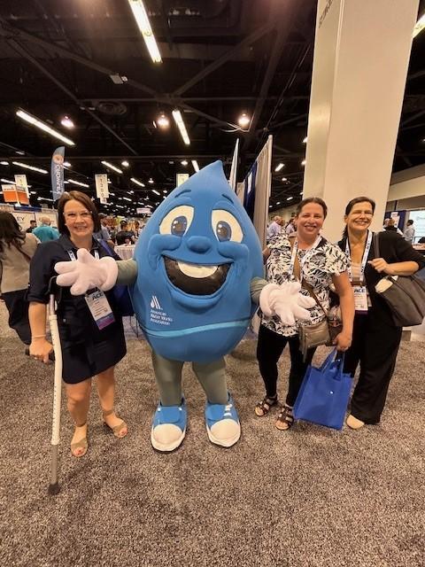 Three women at a conference stand with a mascot dressed like a giant blue water drop with large eyeballs and a toothy smile.