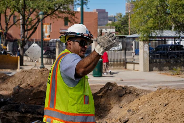 A man in a Denver Water safety vest and hard hat waving traffic forward at a construction site. 