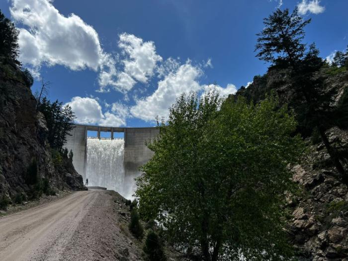 A dirt road leads to a dam with water pour from its outlet structure.