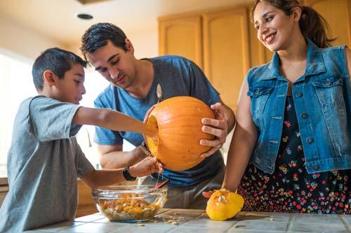 Boy empties pumpkin guts into a bowl while father holds pumpkin. Mother smiles as she looks on at kitchen counter.