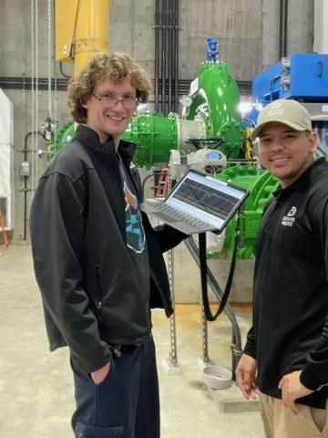 Two men stand with a laptop inside a concrete room with large green pipes.