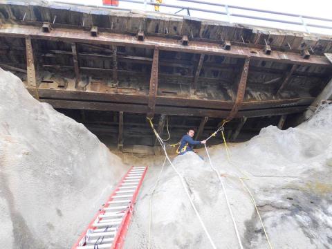 A man is tethered to steel supports and sits on a rocky outcrop to perform a safety inspection.