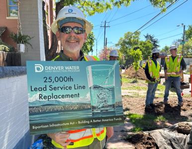 A man in a hard hat, safety vest and sunglasses smiles while holding a sign that reads: Denver Water, 25,000th Lead Service LIne Replaced. In the background are two other construction workers and the edge of a home. 