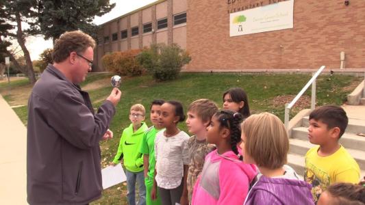 A group of school children watch a man talk, while he holds up a shower head in one hand.