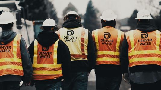 A line of men in Denver WAter safety vests and hard hats, walking away from the camera.