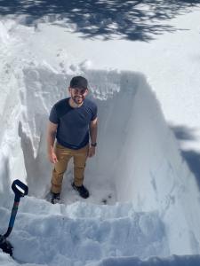 A man stands at the bottom of a pit carved out of snow, smiling up at the camera. The pit is about as deep as he is tall.