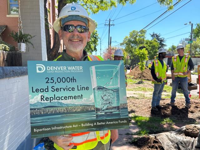 A man in a hard hat, safety vest and sunglasses smiles while holding a sign that reads: Denver Water, 25,000th Lead Service LIne Replaced. In the background are two other construction workers and the edge of a home. 