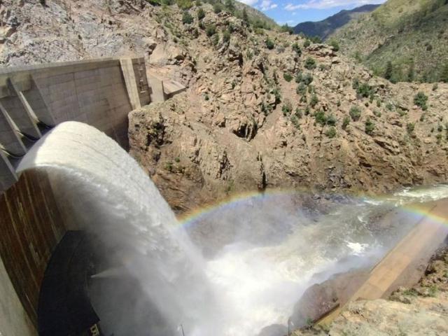Water spills over a dam, arching and crashing into the stream below while sunlight creates a rainbow that dances in the nearby rocks. 