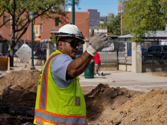 A worker in a yellow safety vest and hard hat, both with the Denver Water logo, waves traffic forward at a construction site.